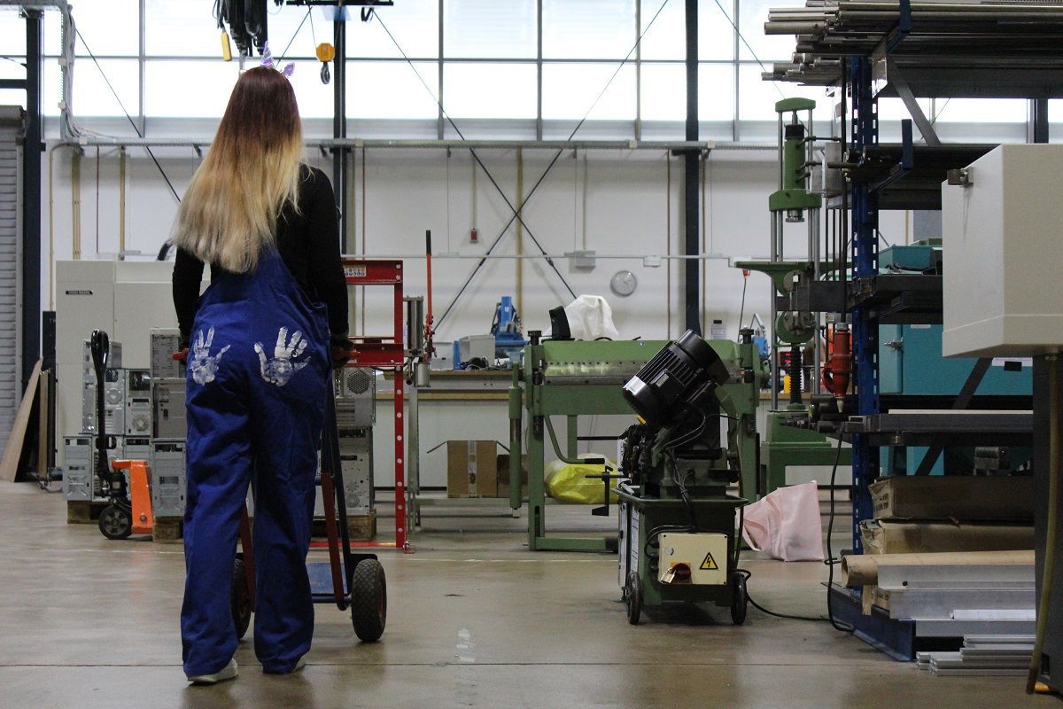 Eine Frau im Blaumann mit zwei weißen Handabdrücken auf dem Po in einer Maschinenhalle.