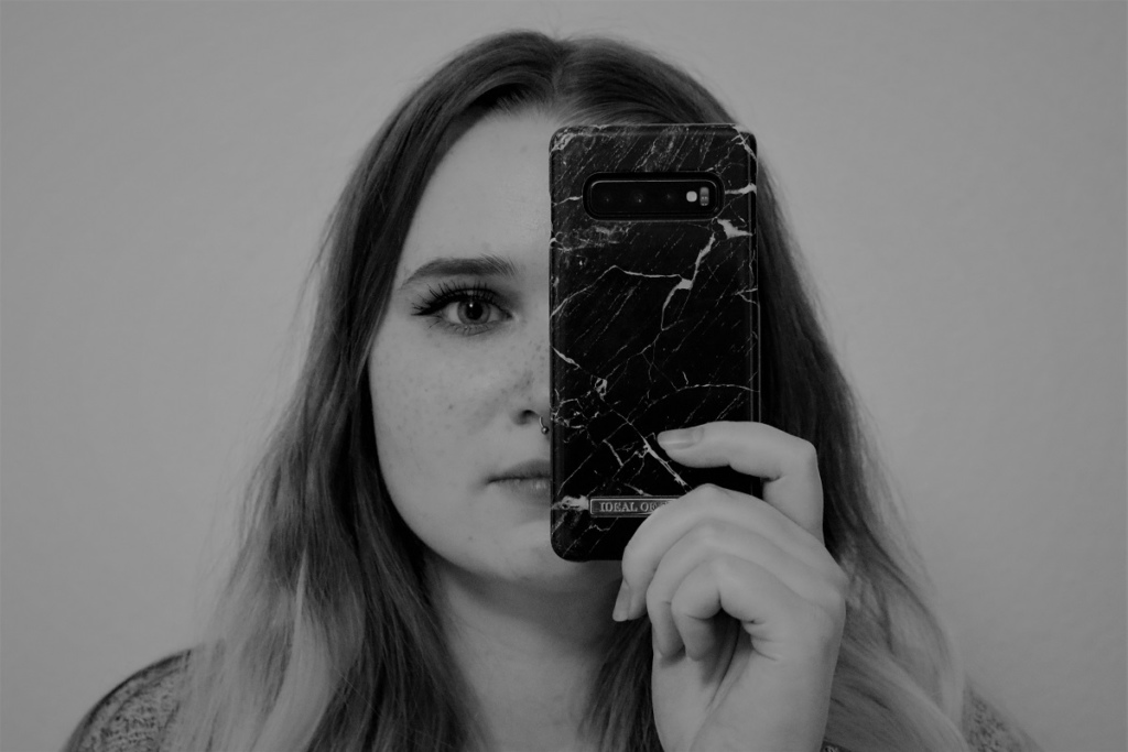 Eine junge Frau, die sich ein Smartphone mit marmorierter Schutzhülle vor die linke Gesichtshälfte hält.