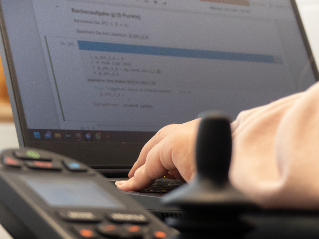 Ein Laptop mit einer Rechenaufgabe im Hintergrund, davor unscharf die Steuereinheit des Rollstuhls