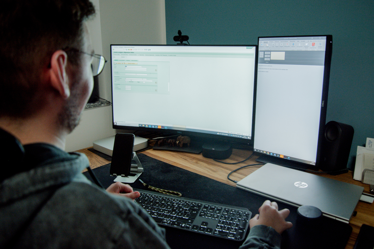Kai an seinem Arbeitsplatz mit zwei Bildschirmen, Smartphone, Laptop, Maus und Tastatur
