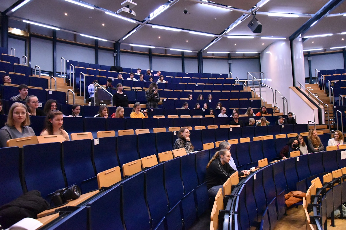 Schüler*innen, Lehrer*innen und Hochschulangehörige sitzen in den Reihen des Hörsaals und folgen der Diskussion